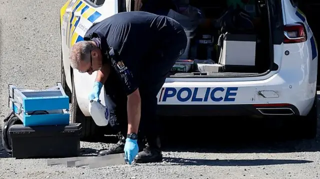 A police office in all navy bends down to pick up a piece of metal on a grey road, he stands in front of his car