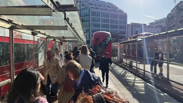People queue up for bus, sun is shining and there are a couple of double decker red busses in sight.