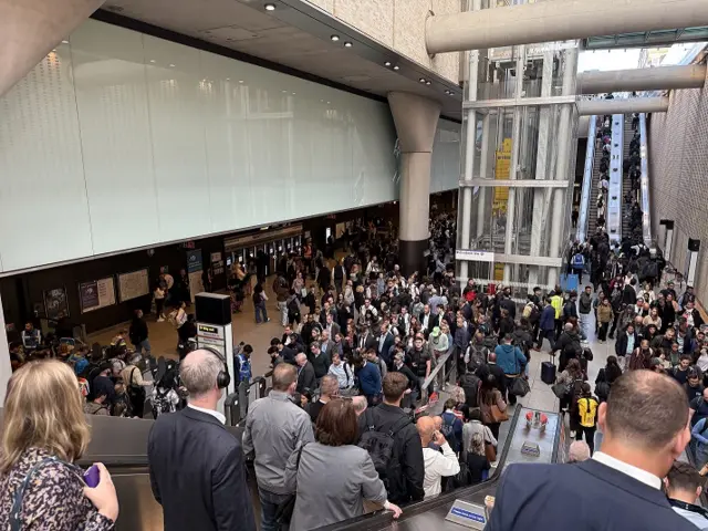 Hundreds of people at a tube station in central London, many wearing suits and work attire. Some going down the escalators, most others congregate near the tube doors.