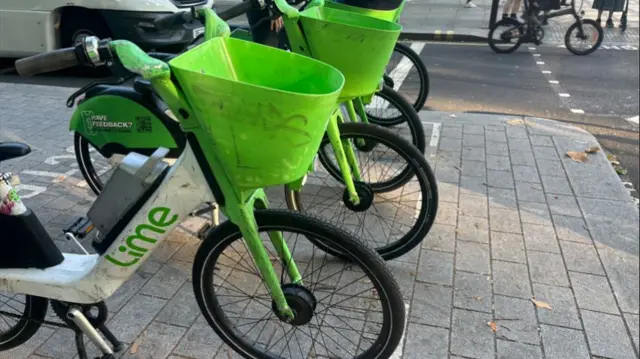 A row of four green bikes which say lime on the side, parked next to a road on a pavement.