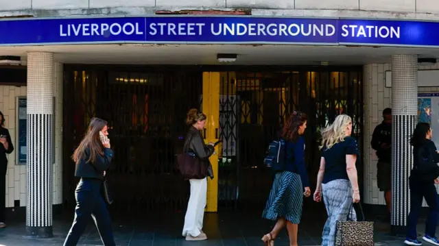 A group of women walking in front of Liverpool Underground Station, which has metal barriers across the closed entrance.