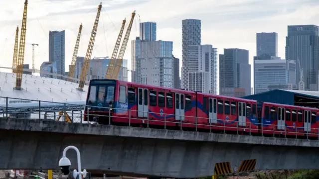 A red Docklands Light Railway train travels past the 02 Arena with the Canary Wharf skyline in the background.