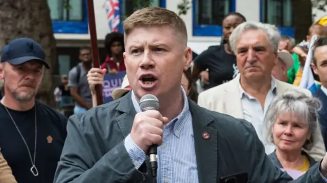 RMT general secretary Eddie Dempsey holding a microphone. He is wearing a light blue shirt and a teal jacket.