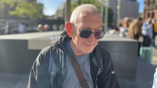 An elderly man wearing a black raincoat and grey shirt looks off to the left. He is wearing black sunglasses but they only half cover his eyes.