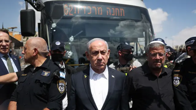 Netanyahu in a black suit and white shirt stands in front of a bus with bullet holes in the wind shield, flanked by emergency workers