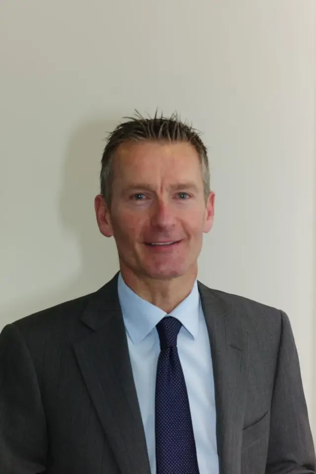Jonathan Hayes, who has grey spiky hair and is wearing a grey suit over a light blue shirt, smiles into the camera