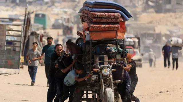 Boys and men either side of a motorbike which pulls a trailer. They push it along with a lot of strength, as the bike is laden several meters high with bedding and wares. They are on a busy dirt road with other vehicles behind them and people walking