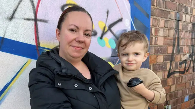 Charlotte Brown-Hughes wearing a black coat holding a one-year-old boy in a brown jumper