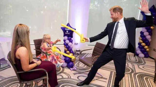 Prince Harry dressed in a dark suit using a long yellow balloon to have a play sword fight with a young girl