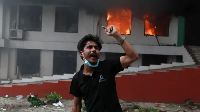 A protester in Nepal wearing black gestures with his arm in front of a burning building