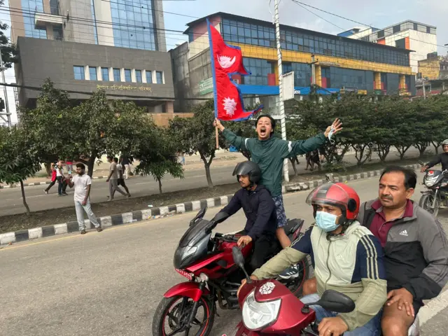 Two motorcycles driving down a road, while a man stands on the back of one, his hands outspread as he holds the flag of Nepal