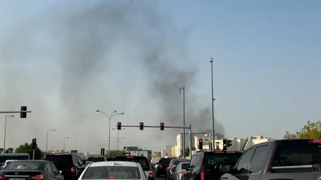 Smoke seen in the sky, while cars wait at traffic lights on a road