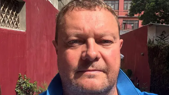 Roger Woodend looks directly at the camera with a stubble beard and thin blonde hair. He is wearing a blue polo shirt with the colour up against the backdrop of colourful red walls