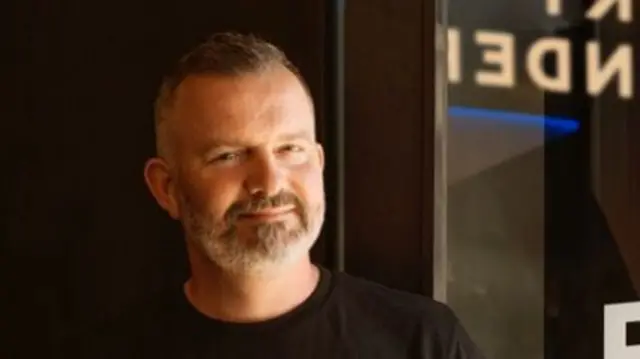 A man with a grey beard and black t-shirt on stands in front of a black wall
