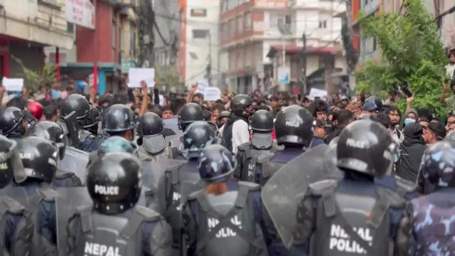 Riot police in a crowd, wearing helmets and cross body protection saying Nepal Police. Crowds of protesters in the distance.