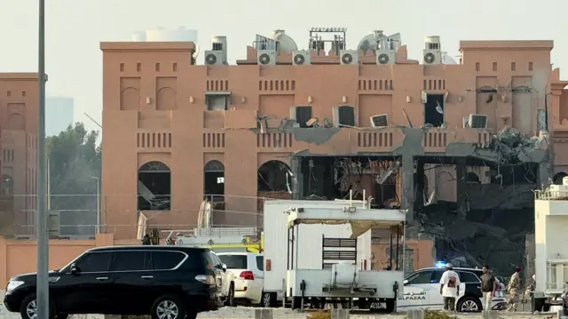 One side of a building with the front right blackened out and destroyed. Some smoke rises and emergency vehicles can be seen out the front