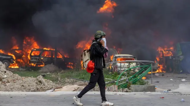 A person walks in front of several cars on fire with black smoke rising, they cover their mouth with a face covering.