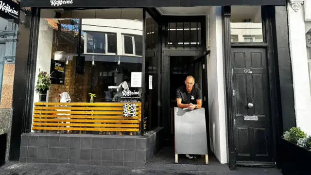 A man stands outside a black painted coffeshop with yellow wooden pannelling