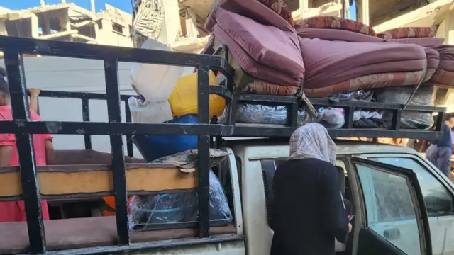 A woman standing next to the open door of a dirty white pick up. She stands facing the car, so the camera only sees the back of her. The car is loaded with bedding, and random wares.