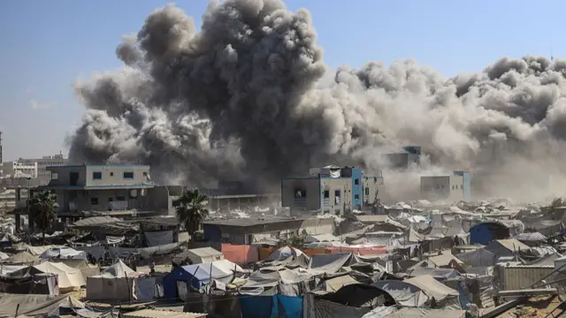 Smoke rising in the background above buildings taking up the whole background of the photo. In front there are hundreds of tents packed closely together