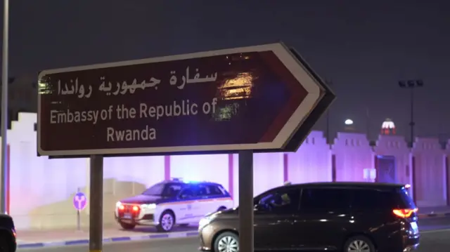 Police car parked near a wall in the background. In the foreground, is a brown arrow-shaped sign pointing right with Embassy of the Republic of Rwanda written on it in Arabic at the top and English at the bottom. Just behind it is a dark coloured car. It is night time.