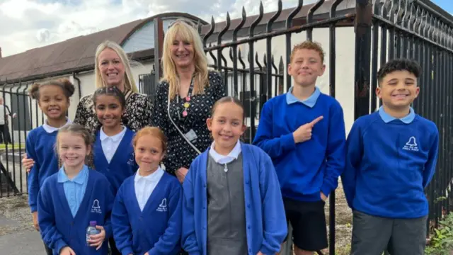 Two teachers stood next to seven pupils dressed in blue school uniform jumpers.
