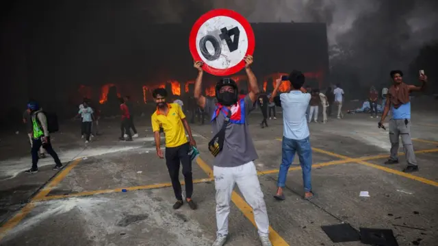 A fire burns inside the Parliament complex during a protest, a man holds a sign saying 40 upside down.