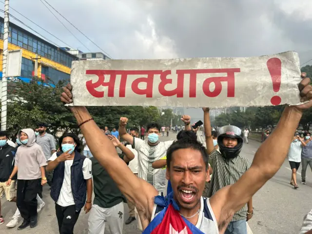 A man yelling in the street holds up a banner saying "attention" in Hindi, while a group of people behind raise their hands in the air triumphantly