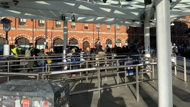 A sunny London street outside of which a queue of people can be seen waiting for taxis