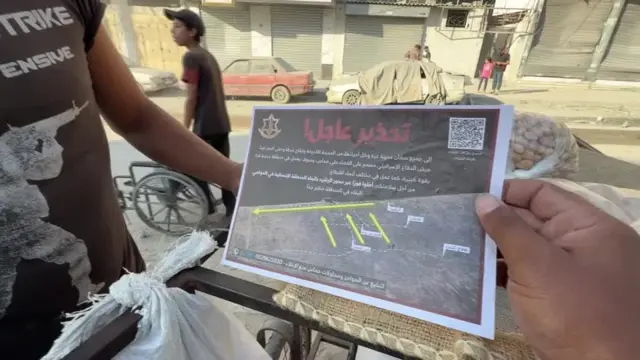 A person holding a leaflet that shows a map with three yellow arrows pointing from inland to the sea, then a line from there along horizontally along the coast. There's big red text in Arabic and writing too