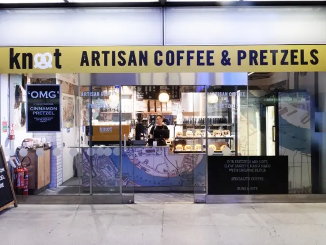 The Knot Artisan Coffee and Pretzels shop at Canary Wharf station. A woman stands behind the counter