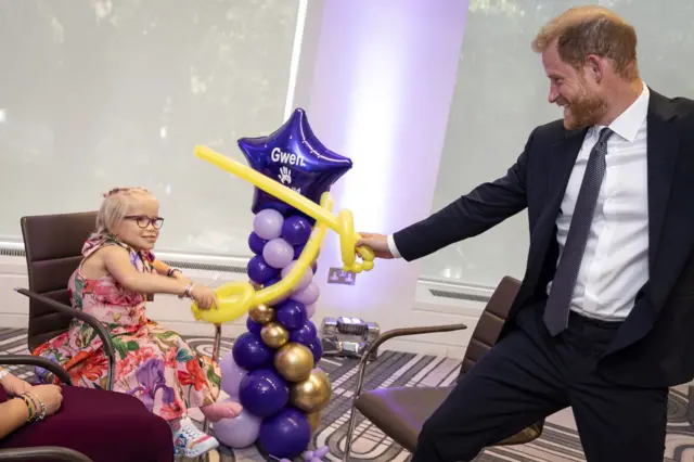 Prince Harry dressed in a dark suit using a long yellow balloon to have a play sword fight with a young girl