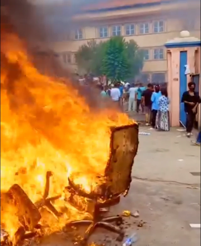 A number of chairs and other debris on fire with protesters walking through the gates of a building in the background