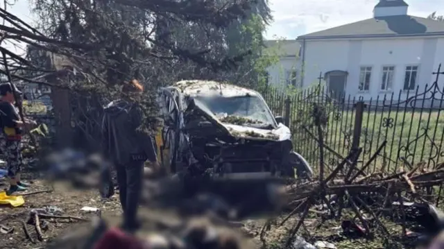 A destroyed car with several blurred out bodies in front of it