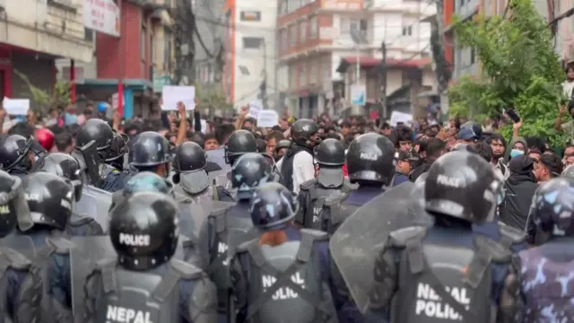 Riot police in the foreground with their backs to the camera and a large crowd of protesters in the background facing the camera