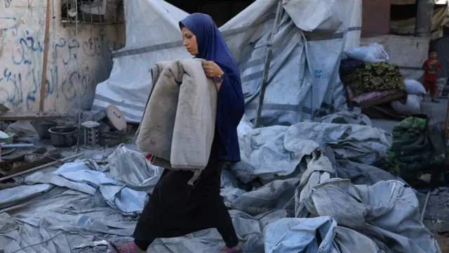 A woman carrying a blanket through rubble and a tent that is falling apart