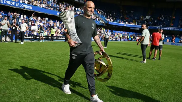 Enzo Maresca carries the Club World Cup trophy and Conference League trophy onto the field at Stamford Bridge
