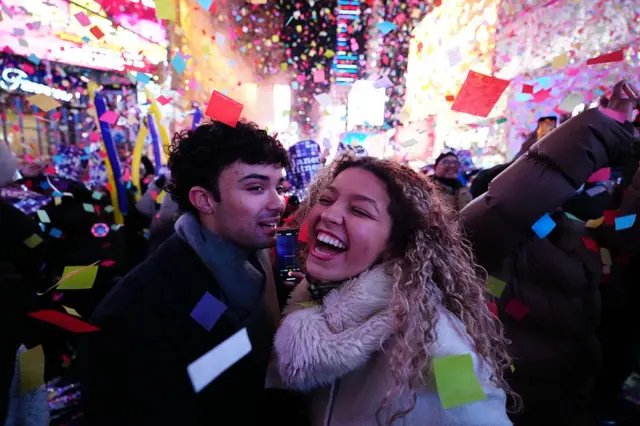 A man and woman with big grins mid celebration at Times Square are surrounded by people and colourful confetti