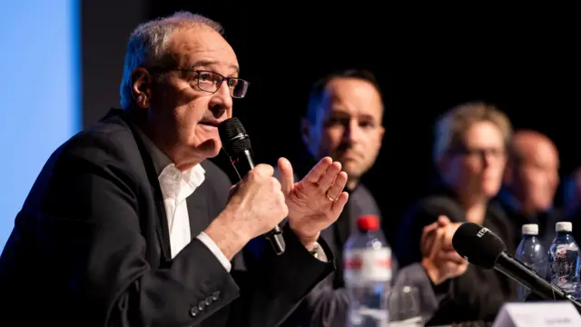 A man in a black suit and white shirt speaks into a microphone at a press conference