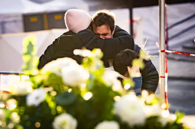 People console one another near the Le Constellation bar. A man is visibly upset. In the foreground are a bunch of white flowers, out of focus.