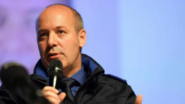 A man with a black coat and blue shirt holds a microphone at a press conference