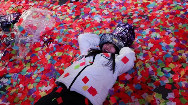 A woman wearing a hat and a white coat lies in multicoloured confetti on the ground in Times Square, New York City