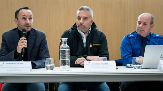 Three officials sit at a table, with the one on the left holding a microphone
