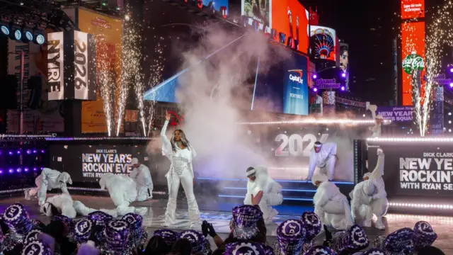 Ciara stands in the middle of a stage dressed in white with her hand pointed to the sky, surrounded by dancers also in white crouching in positions