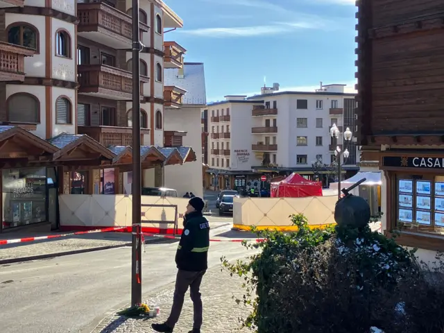 A police officer stands next to a cordoned off area in Crans-Montana, several white sheets and a red plastic forensic tent surround the centre of a square outside Le Constellation bar