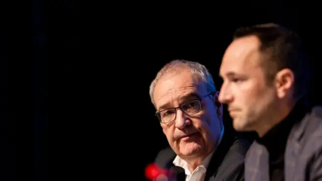 Two men sit at a table holding a press conference