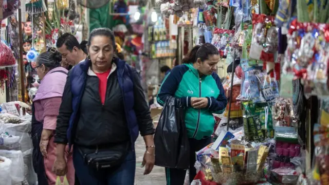 A woman walks out of focus at the front of an image with a couple shopping behind in a market, with a woman in a green jacket on the right looking at goods.
