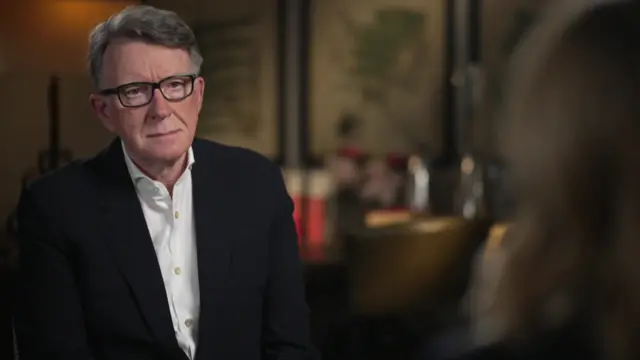 Lord Peter Mandelson sits looking at Laura Kuenssberg during an interview. He has short, grey hair, glasses, and is wearing a black jacket and white shirt.