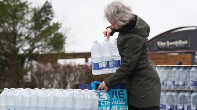 A woman wearing a green coat lifts a six-pack of water into a reusable bag at a water point. We can see lots of bottles of water behind her