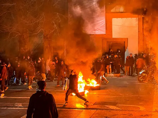 Protesters gather in a street in Iran with one person seen standing in front of a tyre set on fire